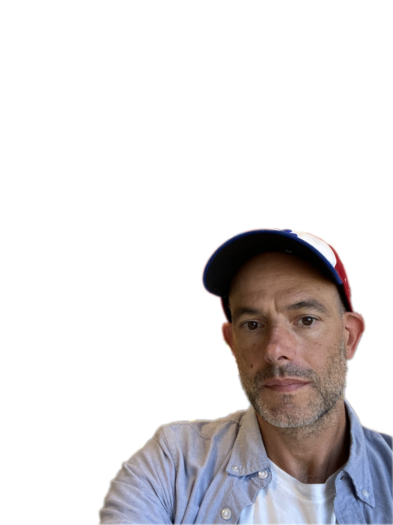 Light-skinned male-seeming figure wearing a baseball cap, a blue collared shirt, and white round-neck tshirt under. The figure looks slightly below the camera, as if in a selfie.