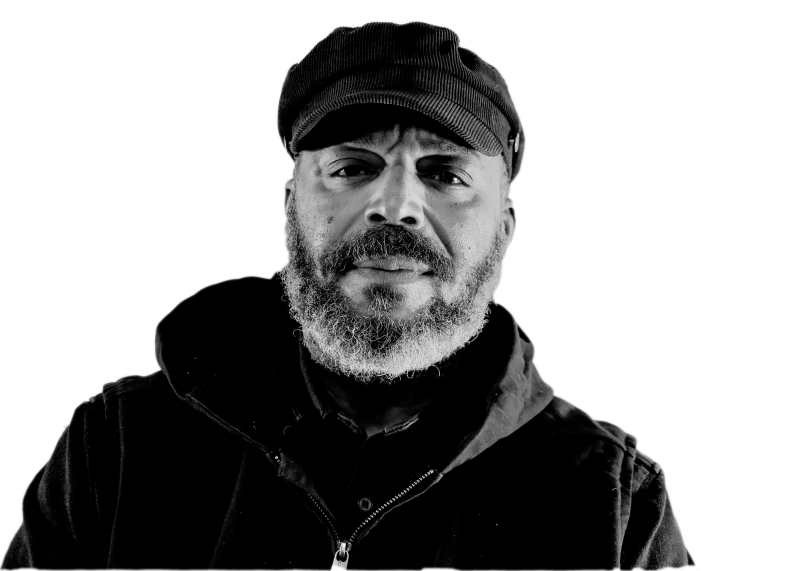 Black and white photograph of a dark-skinned figure with a beard and black baseball cap looks directly into the camera. The person wears a black zip up jacket.