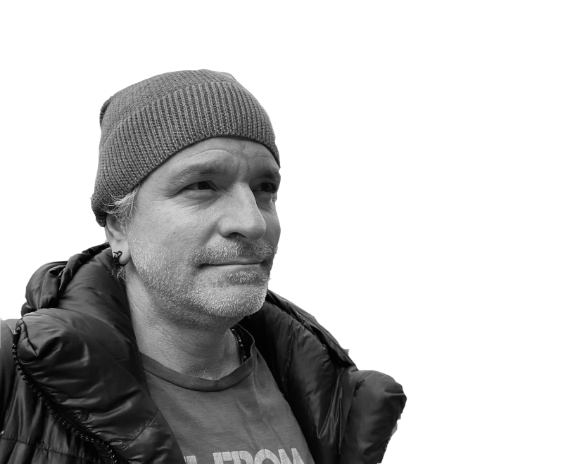 Black and white photograph of male seeming figure with a beany cap, earring, and slight stubble, wearing a t-shirt and a puffy winter coat.